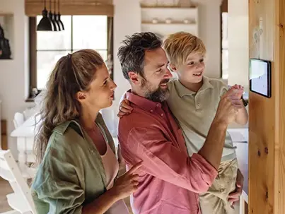 A family, with a man holding a child and a woman, interacts with a smart home tablet
