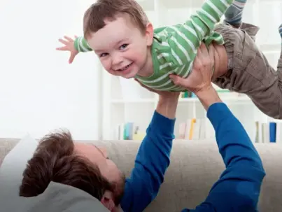 A joyful father lies on a couch, lifting his smiling young son