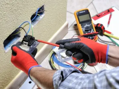 An electrician in red gloves works on a wall electrical outlet