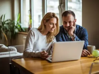 A smiling couple sits at a wooden table, looking at a laptop together
