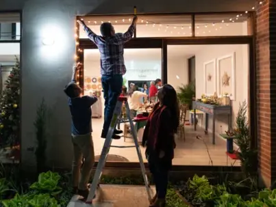 Three people decorate a house exterior with string lights