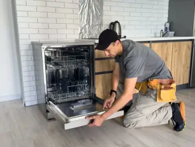 A technician in a black cap and tool belt kneels to examine an open dishwasher in a modern kitchen