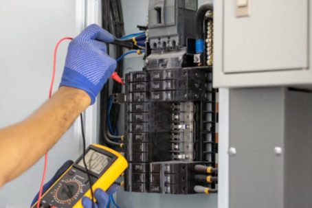An electrician in blue gloves uses a yellow multimeter to test an open electrical panel